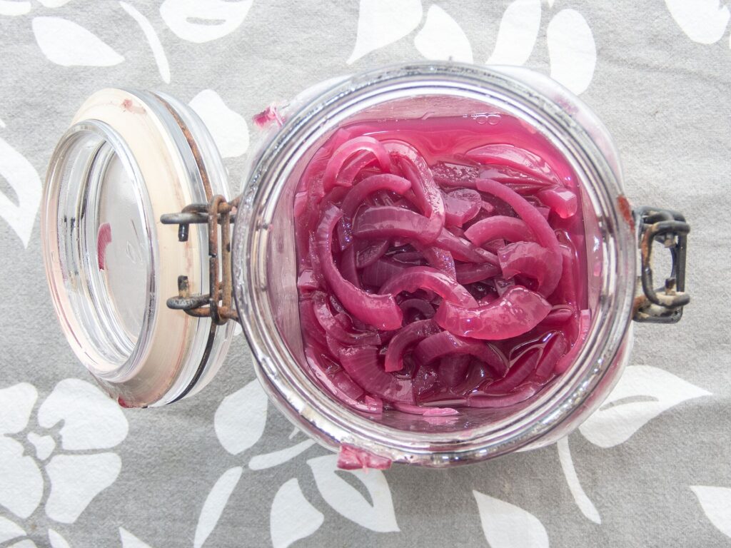 A glass jar of thinly sliced pickled red onions standing open beside a spoon