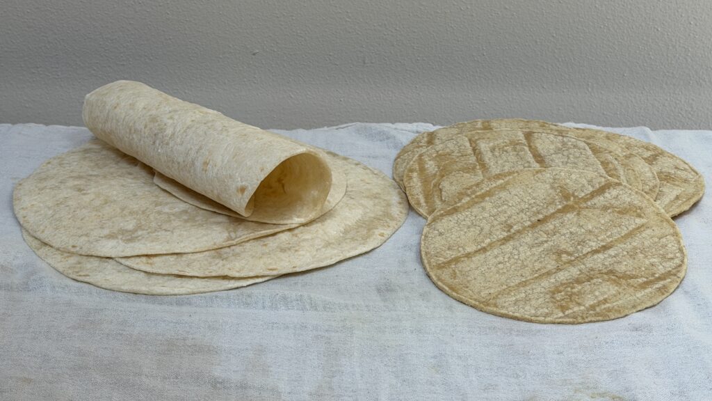 A stack of soft flour tortillas on a wooden board next to a folded cloth