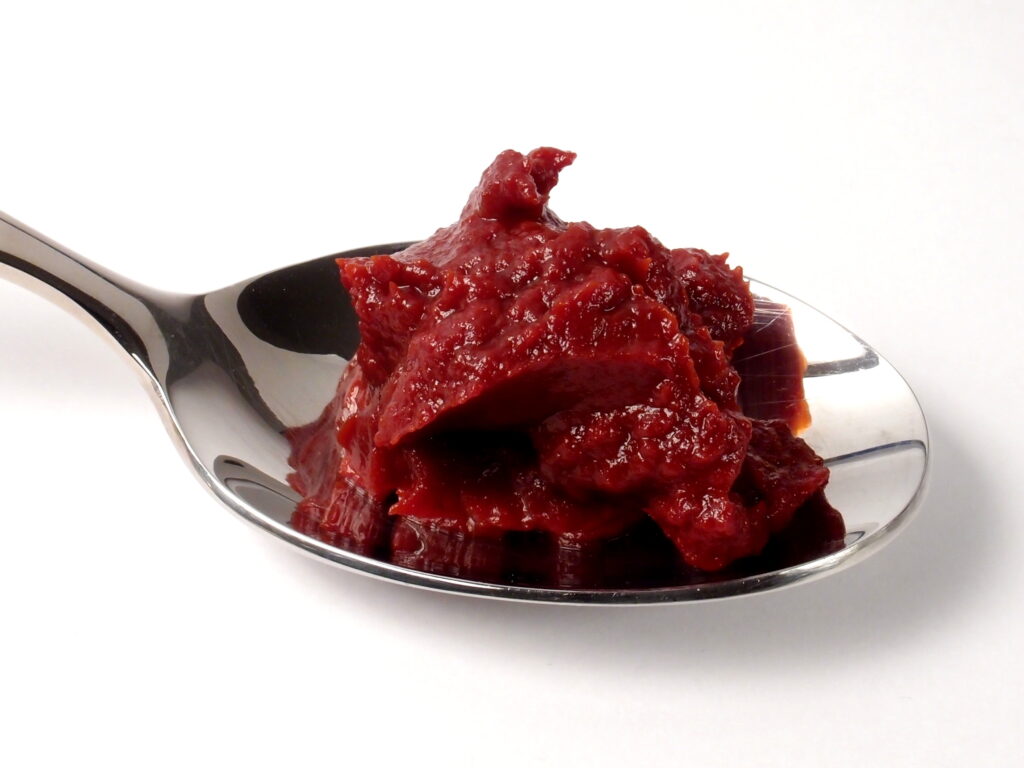 Dark red tomato paste on a spoon above an open can on a counter